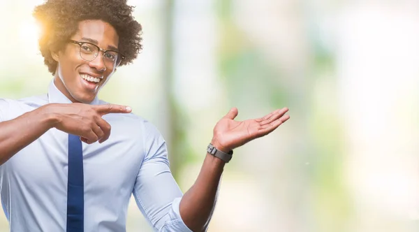Afro american business man wearing glasses over isolated background amazed and smiling to the camera while presenting with hand and pointing with finger.