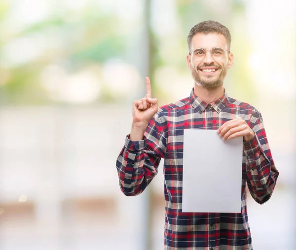 年轻的时髦的成人男子持有空白纸表惊讶与一个想法或问题指向手指与幸福的脸 — 图库照片