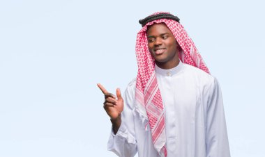 Kameraya bakarak yan el ve parmak ile işaret eden geleneksel keffiyeh yüzünde kocaman bir gülümseme ile izole arka plan üzerinde giyen genç Arapça Afrika adam.