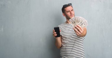 Beyaz delikanlı yüz çapraz silah ile dolar ve smarphone şüpheci ve sinir, onaylamayan ifade tutan gri grunge duvarın üzerinden. Negatif kişi.