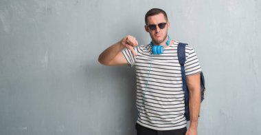 Gri grunge duvar kulaklık ve sırt çantası kızgın yüzü, eksi işareti aşağı antipati başparmak ile gösterilen, ret kavramı ile giyen genç beyaz öğrenci adam