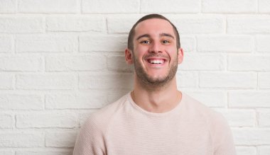 Mutlu yüz ayakta beyaz tuğla duvar üzerinde duran ve diş gösterilen kendine güvenen bir gülümsemeyle gülümseyerek beyaz delikanlı