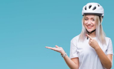 Hayret ve el ile sunulması ve parmak ile işaret ederken kameraya gülümseyen izole arka plan üzerinde bisikletçi Güvenlik kask giyen genç sarışın kadın.