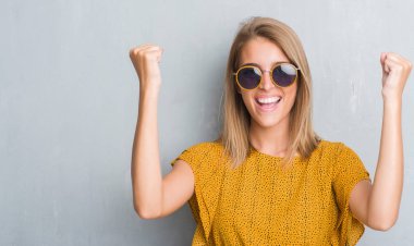 Duygu tezahürat gurur çığlık ve zafer ve başarı çok heyecanlı, kutluyor retro güneş gözlüğü takıyor grunge gri duvar üzerinde duran güzel genç kadın