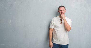 Beyaz delikanlı emin çapraz kollar ve çeneme kaldırdı el ile gülümseme ile kameraya bakarak güneş gözlüğü takıyor gri grunge duvarın üzerinden. Pozitif düşünce.