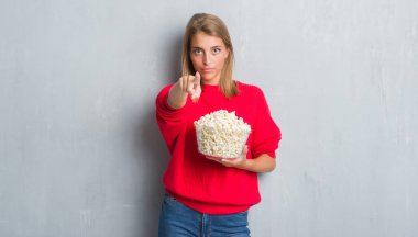 Güzel genç kadın kamera ve size parmak ile işaret pop Mısır yeme grunge gri duvarın üzerinden, el işareti, pozitif ve kendinden emin bir jest ön