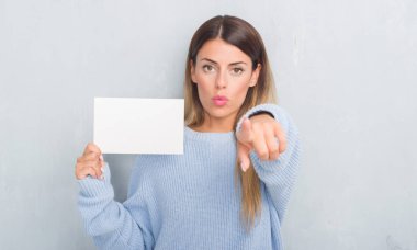 Genç Yetişkin kadın boş kartı kamera ve size parmak ile işaret tutan gri grunge duvarın üzerinden, el işareti, pozitif ve kendinden emin bir jest ön