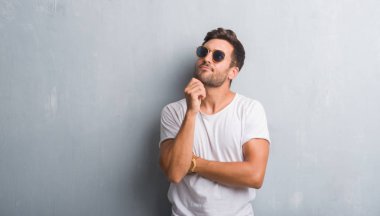 Çene düşünme hakkında soru, dalgın ifade üzerine el ile güneş gözlüğü takıyor gri grunge duvarın üzerinden yakışıklı genç adam. Düşünceli yüzüyle gülümsüyor. Şüphe kavramı.