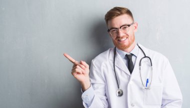 Genç redhead Doktor adam gri grunge duvar el ve parmak ile tarafına işaret çok mutlu