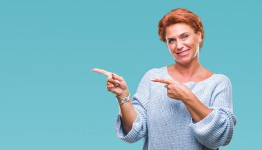 Gülümseyen ve iki elleri ve parmakları ile tarafına işaret kamera bakarak izole arka plan üzerinde kış kazak giyen Atrractive üst düzey beyaz Kızıl saçlı kadın.