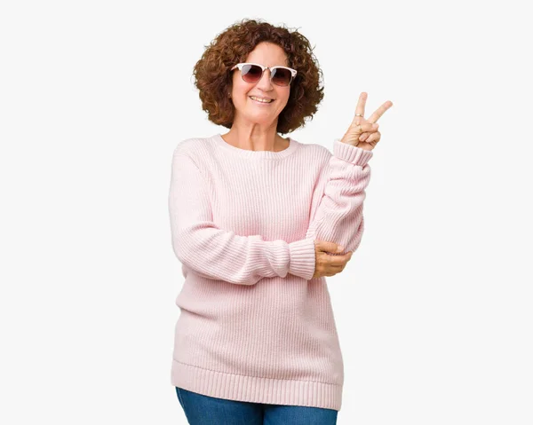 美丽的中间位高级妇女穿着粉红色的毛衣和太阳镜在孤立的背景微笑着与幸福的脸眨眼对着相机做胜利的标志 — 图库照片