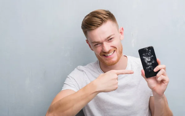 Young redhead man over grey grunge wall holding broken smartphone very happy pointing with hand and finger