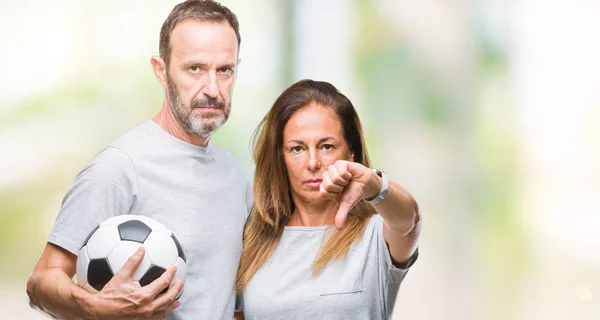 Middle age hispanic couple holding football soccer ball over isolated background with angry face, negative sign showing dislike with thumbs down, rejection concept