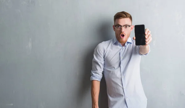 Young redhead man over grey grunge wall showing blank screen of smartphone scared in shock with a surprise face, afraid and excited with fear expression