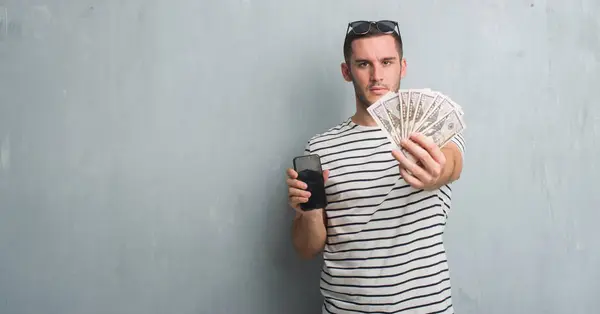 Young Caucasian Man Grey Grunge Wall Holding Dollars Smarphone Serious — Stock Photo, Image
