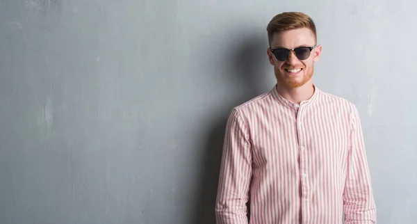 Young redhead man over grey grunge wall wearing sunglasses with a happy face standing and smiling with a confident smile showing teeth