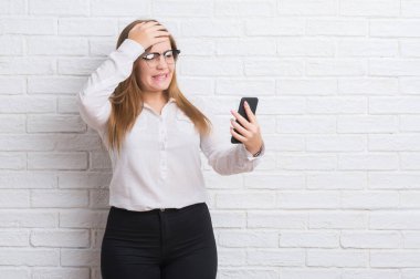 Genç Yetişkin iş kadını kafasına, el ile vurguladı smartphone kullanarak ileti gönderme beyaz tuğla duvarın üzerinden şok utanç ve sürpriz suratla, öfkeli ve sinirli. Korku ve hata için üzgün.