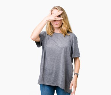 Mahcup ifadeyle parmaklarımın arasından seyir yüz ve gözler el ile kapsayan şok bakmak izole arka plan üzerinde büyük boy rahat t-shirt giyen güzel genç kadın.
