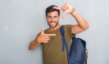 Üzerinde gri grunge duvar giyen genç adam yakışıklı turist sırt çantası elle yapma çerçeve ve parmakları ile mutlu yüz gülümseyerek. Yaratıcılık ve Fotoğrafçılık kavramı.