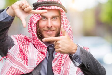 Genç yakışıklı Arap adam uzun saçlı elle yapma çerçeve ve parmakları ile mutlu yüz gülümseyen izole arka plan üzerinde keffiyeh giyiyor. Yaratıcılık ve Fotoğrafçılık kavramı.