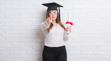 Genç Yetişkin kadın beyaz tuğla duvar derecesi kamera ve size parmak ile işaret tutan lisansüstü şapkası yüzünden el işareti, pozitif ve kendinden emin bir jest ön