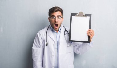 Yakışıklı genç adam gri grunge duvar holding Pano şok sürpriz yüzü, korkuyor ve korku ifadesi ile heyecanlı olan korkmuş üzerinde doktor