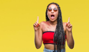 Hayret ve yukarıya bakıyor ve parmakları ile işaret ve silah kaldırdı sürpriz izole arka plan üzerinde güneş gözlüğü takıyor Doğum işareti ile saç Afrikalı-Amerikalı genç örgülü.