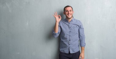 Üzerinde gri grunge duvar gülümseyen Genç beyaz adam el ve parmaklar ok işaretiyle yapıyor olumlu. Başarılı ifade.