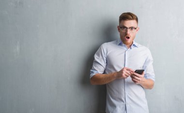 Genç Kızıl saçlı adam gri grunge duvar bir sürpriz yüz, korkuyor ve heyecanlı korku ifadesi ile smartphone şokta korktuğunu kullanarak ileti gönderme