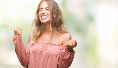 Gülümseyen ve başarı için çığlık güzel sarışın genç kadın üzerinde izole arka plan çok mutlu ve heyecanlı kaldırdı, silah ile kazanan jest yapıyor. Kutlama kavramı.