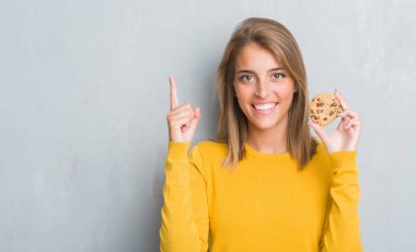 Güzel genç kadın çikolata yeme grunge gri duvarın üzerinden bisküvi bir fikirle sürpriz çip ya da gülen yüz, bir numara ile işaret parmağı soru