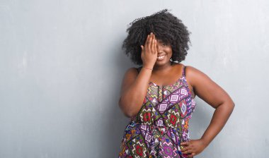 Gri grunge duvar renkli giyen genç Afro-Amerikan kadın elbise yüz ve sürpriz duygu kendinden emin gülümseme ile el ile bir göz kapsayan.