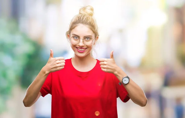 Young beautiful blonde woman wearing red t-shirt and glasses over isolated background success sign doing positive gesture with hand, thumbs up smiling and happy. Looking at the camera with cheerful expression, winner gesture.