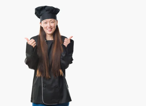年轻的中国女人在孤立的背景穿着厨师制服的成功标志做积极的手势用手 竖起大拇指微笑和快乐 带着欢快的表情 获胜者的手势看着镜头 — 图库照片