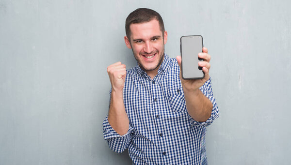 Молодой кавказский мужчина над серой гранж-стеной показывает чистый экран смартфона, крича гордо и празднуя победу и успех очень взволнован, аплодируя эмоциям
