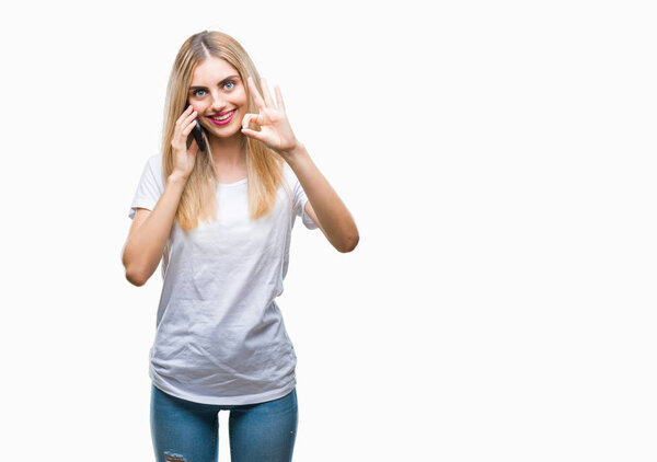 Young beautiful blonde woman talking using smartphone over isolated background doing ok sign with fingers, excellent symbol