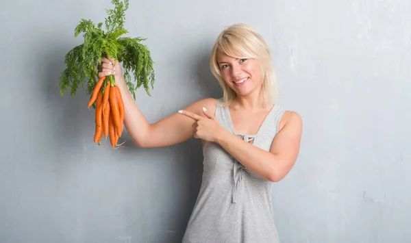 白种人成年妇女在灰色的格格墙举行新鲜的胡萝卜非常高兴地指向用手和手指 — 图库照片