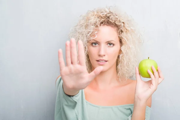 年轻的金发碧眼的女人在格格灰色背景吃绿色苹果与开放的手做停止标志用严肃和自信的表情 防御姿态 — 图库照片