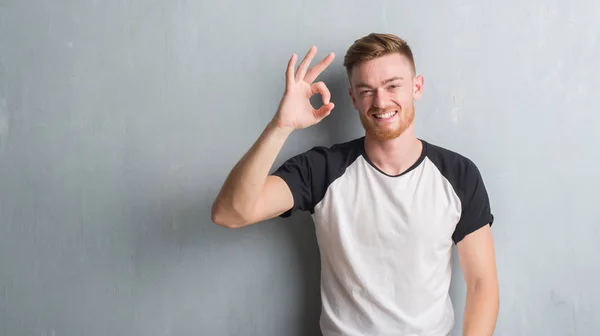 年轻的红发男子在灰色的格格墙微笑积极做确定的标志与手和手指 成功的表达 — 图库照片