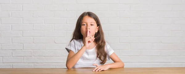 自宅のテーブルの上に座って若いヒスパニックの子供は 唇に指で静かにするように求めています 沈黙と秘密の概念 — ストック写真