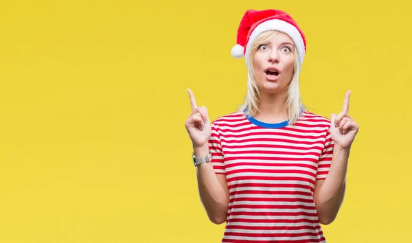 年轻美丽的金发女子戴着圣诞节帽子在孤立的背景惊讶和惊讶抬起手指和举起的胳膊 — 图库照片