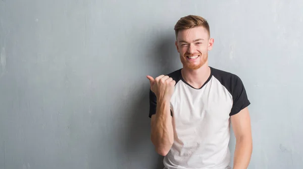 Young redhead man over grey grunge wall smiling with happy face looking and pointing to the side with thumb up.