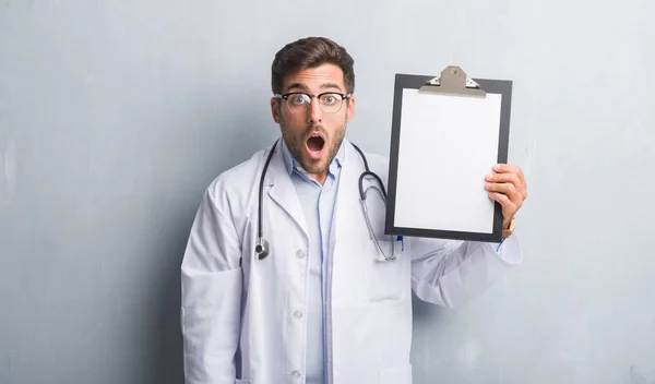 Handsome Young Doctor Man Grey Grunge Wall Holding Clipboard Scared — Stock Photo, Image