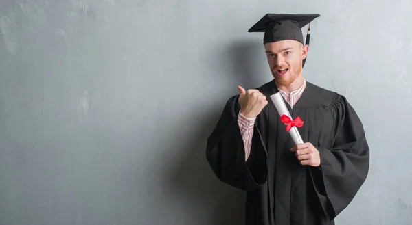 灰色のグランジの赤毛の若い男壁の身に着けている大学院制服指していると幸せそうな顔を笑顔で側まで親指で示すの学位を保持 — ストック写真