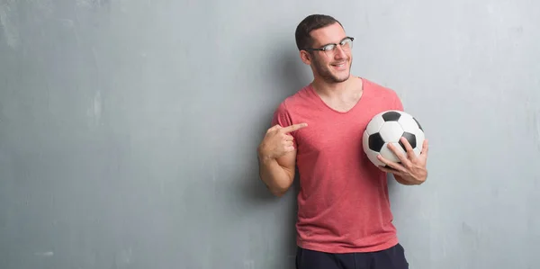 サッカー サッカー ボールを手や指で指している非常に満足して保持している灰色のグランジの壁を越えて若い白人男 — ストック写真