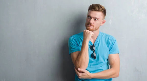 Young redhead man over grey grunge wall wearing casual outfit with hand on chin thinking about question, pensive expression. Smiling with thoughtful face. Doubt concept.