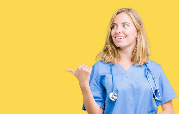 美丽的年轻医生妇女穿着医疗制服在孤立的背景微笑与愉快的面孔看和指向边用拇指 — 图库照片