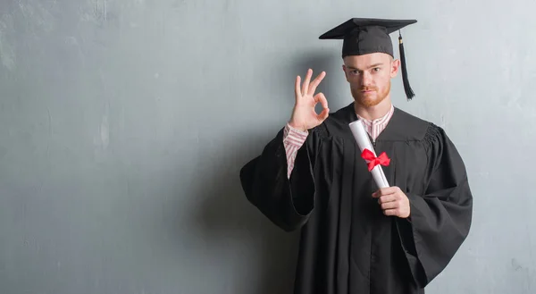 灰色のグランジの赤毛の若い男壁の身に着けている大学院制服優秀なシンボルの指で サインをして学位を保持 — ストック写真