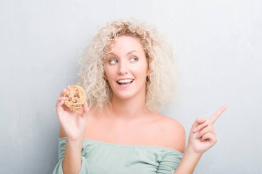 Sarışın genç kadın grunge üzerinde gri duvar çikolatalı bisküvi el ve parmak ile tarafına işaret çok mutlu yemek