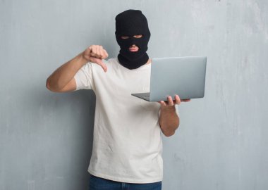 Genç beyaz hacker adam dizüstü kullanarak kızgın yüzü, eksi işareti aşağı antipati başparmak ile gösterilen, ret kavramı ile siber saldırı yapmak gri grunge duvarın üzerinden